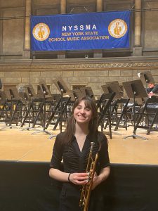 VCHS senior Amelia K holding a trumpet with a banner behind her that says NYSSMA New York State School Music Association.