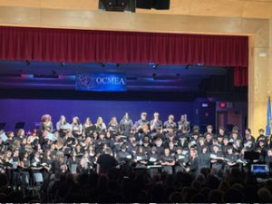 The Chorus at the OCMEA Senior High All-County Music Festival is performing on stage. The students are singing and looking down at their music. They are all wearing black and there are four layers of students on risers singing to the crowd. 