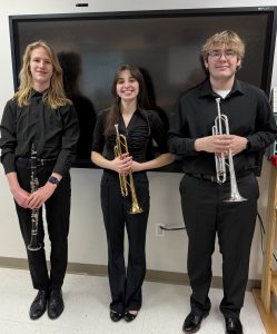 Three students smile for the camera while holding their instruments and wearing all black. The student on the left is holding a clarinet, in the middle and on the right the students are holding trumpets. 