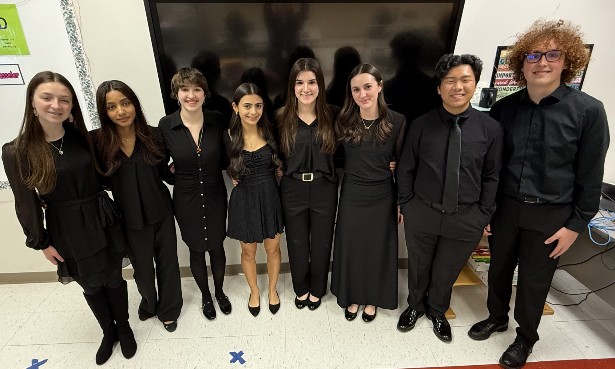 A group of student musicians stand together wearing black and pose. There are eight students total and they are in a line smiling for the camera at the OCMEA Senior High All-County Music Festival.