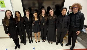 A group of student musicians stand together wearing black and pose. There are eight students total and they are in a line smiling for the camera at the OCMEA Senior High All-County Music Festival. 