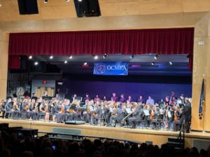 A wide shot of the students on stage performing at the OCMEA Senior High All-County Music Festival. The entire stage is in the picture, with the wall in front of the stage also shown. All the students are wearing black and playing their instruments. 