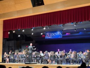 The band is performing at the OCMEA Senior High All-County Music Festival. The students are all sitting down and playing their instruments in front of sheet music with a conductor facing them. 