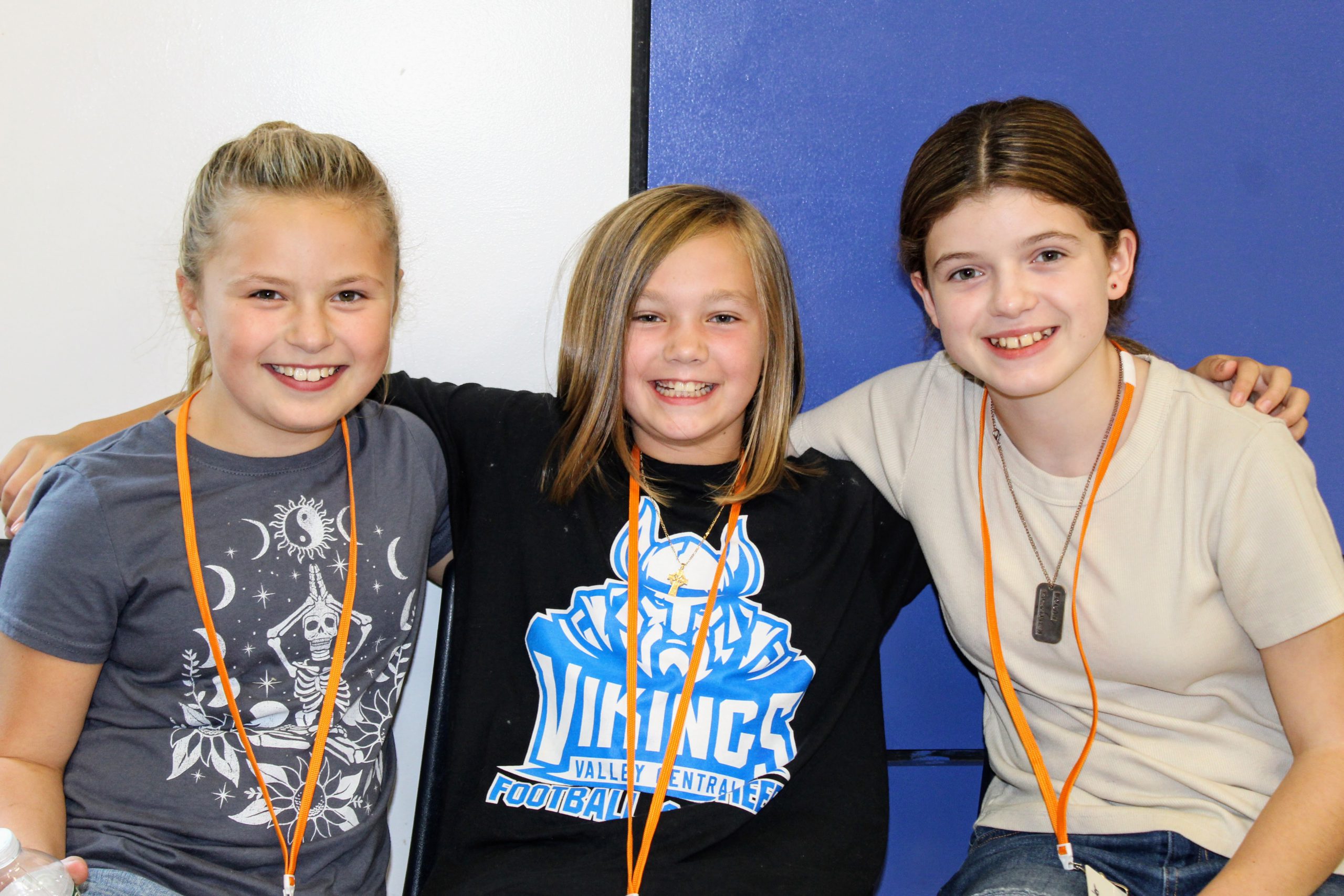 Three students are sitting together and smiling at the camera in front of Valley Central colors on the wall. 